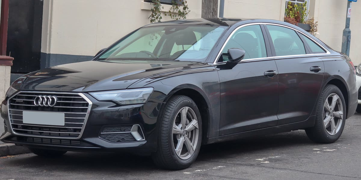 Original front-corner photograph of a dark Audi parked beside a building, with reflections across its hood and doors
