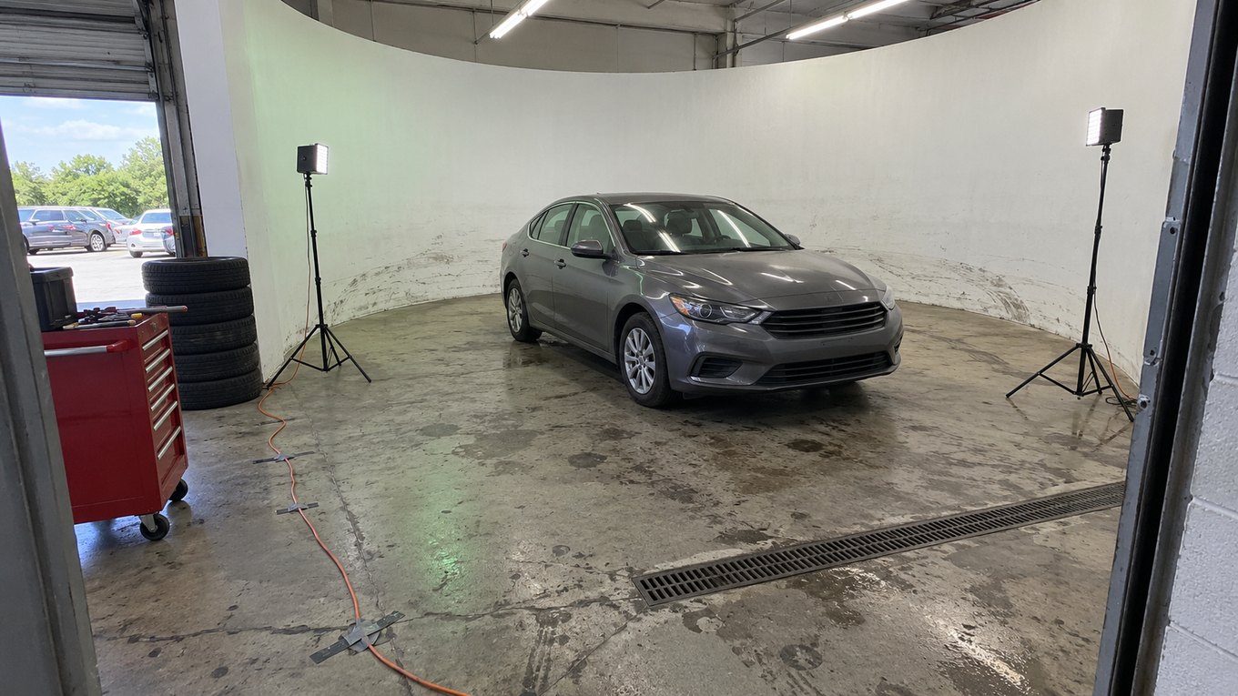 A service bay converted into a car photo booth, with a scuffed white curved wall, two LED lights on stands and a gray sedan in the middle