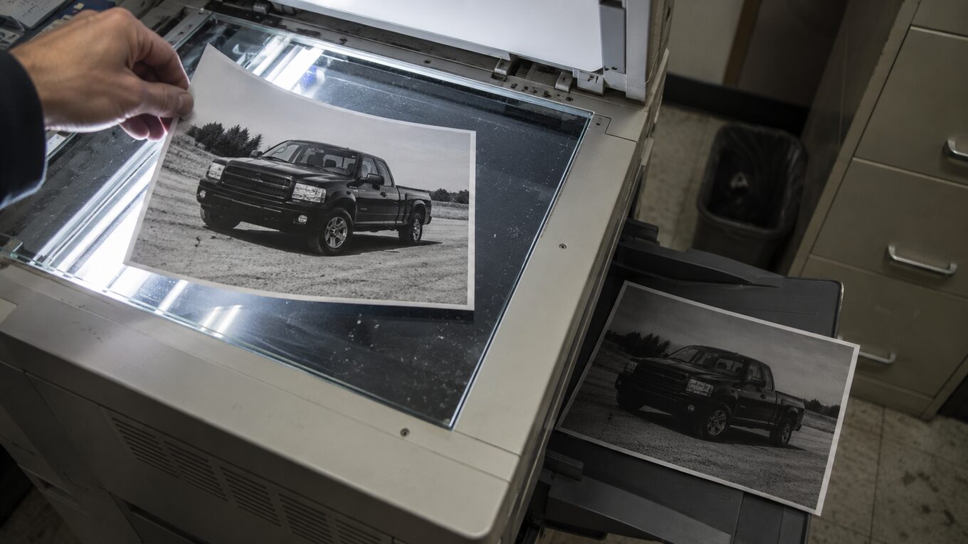 A printed vehicle photo being lifted off a photocopier glass while a second, darker copy of it sits in the output tray