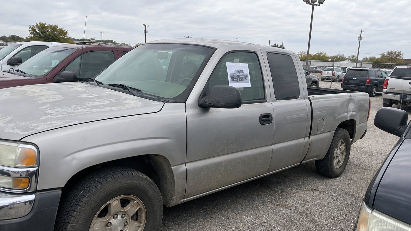 A dusty silver used pickup on a gravel dealer lot with a printed brochure photo of a new truck taped to its window